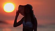 Woman in white bikini top, straw sun hat on beach at sunrise during weekend holiday at golden hour