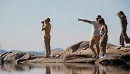 Group of stylish young people walking on rocky beach and enjoying time in nature, their friend taking photograph of nature