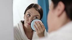 A woman looking at herself in the mirror applies powder with a soft puff. The girl admires herself and removes makeup with a reusable sponge.