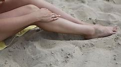 Woman Applying Sunscreen On Her Legs At Beach Whilst Sitting On Yellow Beach Towel
