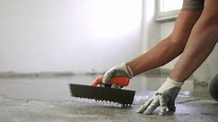 Workman pouring self-leveling screed on floor, spreading it and smoothing with metal spatula standing on his knees, building master aligning fresh floor surface on construction site. Concrete mixture