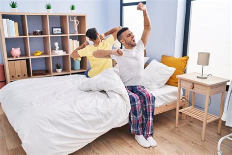 Two Man Couple Waking Up Stretching Arms Sitting On Bed At Bedroom