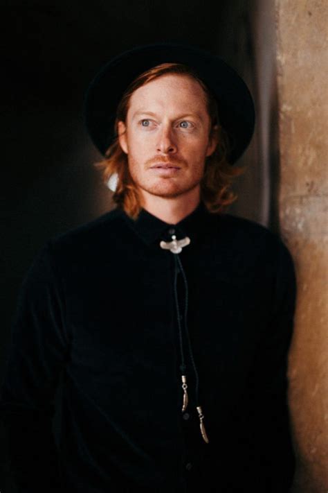 A Man With Long Hair Wearing A Black Shirt And Hat Standing Next To A Wall