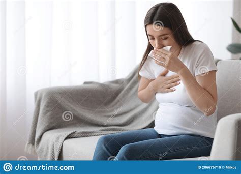 Sick Pregnant Woman Sitting On Couch And Coughing Into Tissue Stock