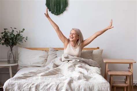 Aged Female Waking Up In Morning Stretches Seated In Bed Stock Image