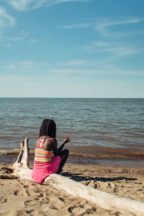 black girl sitting on a log by the beach by stocksy contributor gabi