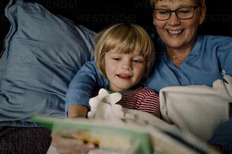 Smiling Grandmother Reading Story Book For Grandson On Bed At Home