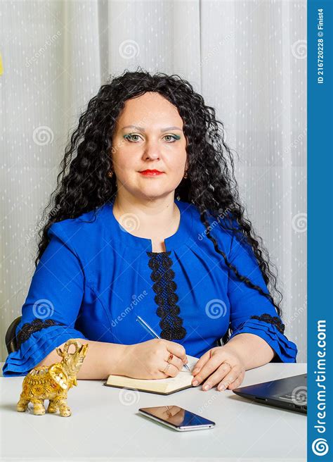 Curly Brunette Woman Sits At The Table In The Office With A Laptop