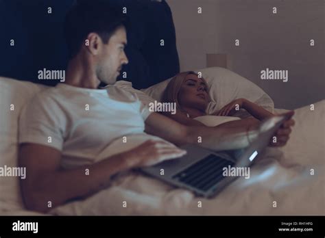 Suspicious Young Man Using Laptop And Looking At Girlfriend Sleeping In