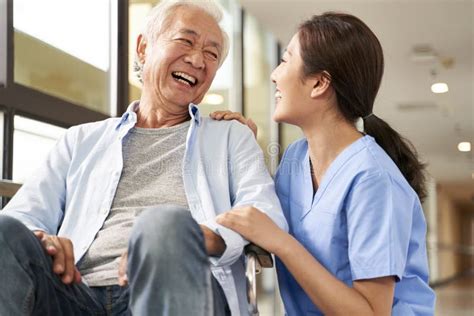 Young Friendly Asian Female Caregiver Talking To Elderly Man In Nursing