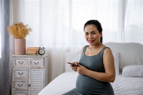 Asian Pregnant Mother Sitting On Bed And Holding Mobile Phone Stock