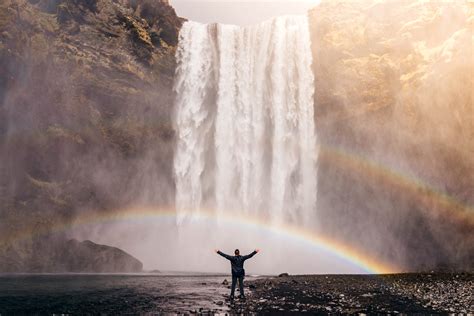 man standing beneath  waterfall  double rainbow image