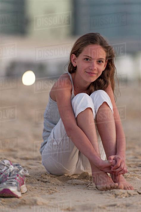 preteen girl sitting on beach with barefeet hugging knees