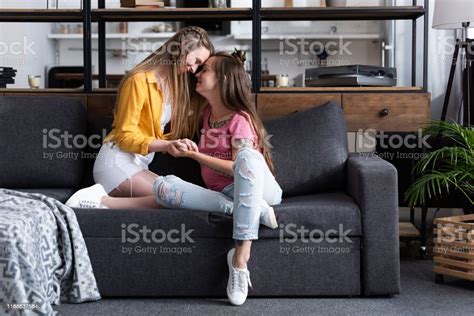 Two Smiling Lesbians Holding Hands While Sitting On Sofa In Living Room