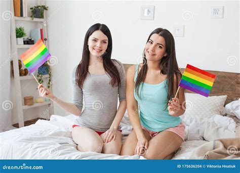 Lesbian Couple In Bedroom At Home Sitting Holding Lgbt Color Flags