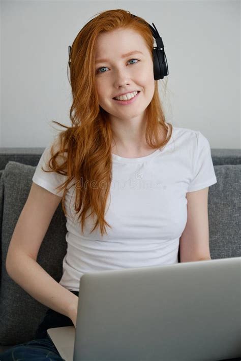 A Cute Redhead Girl Sits On A Sofa In A White T Shirt And Headphones