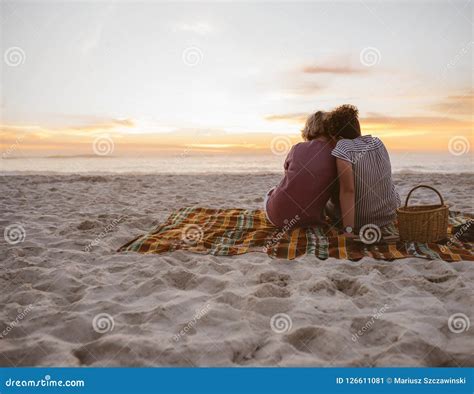 Young Lesbian Couple Sitting On Beach Blanket Watching The Sunset Stock