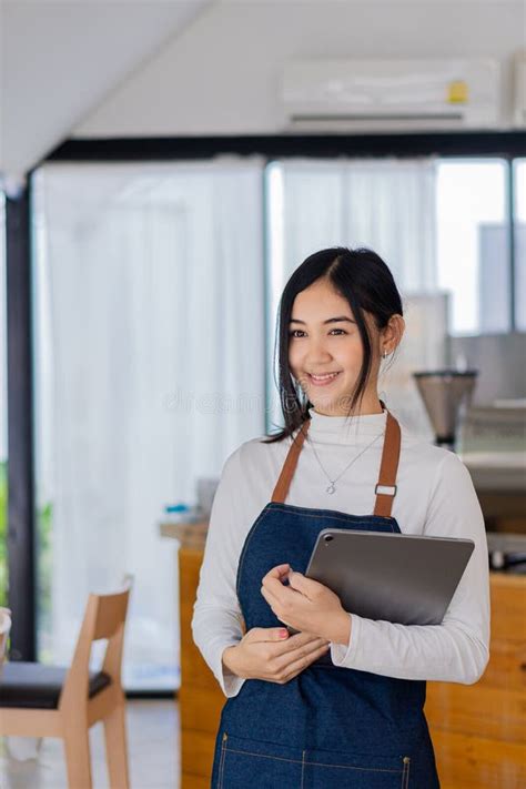 Pretty Asian Barista In Apron Holding A Tablet And Standing In Front Of