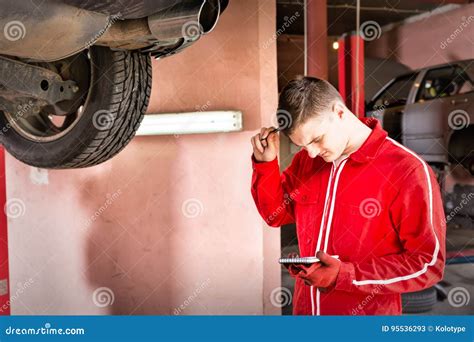 young confused male motor mechanic standing making notes   lifted car stock image