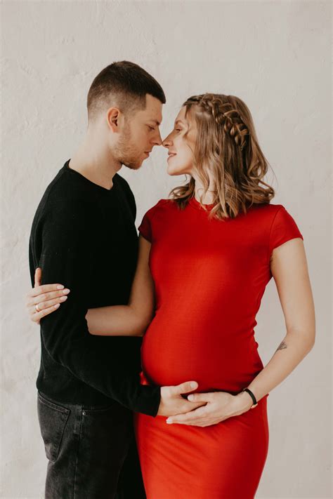 A Pregnant Woman In A Red Dress Standing Next To A Man With His Hand On