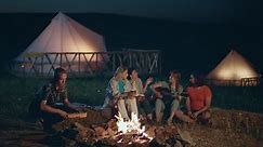 In front of the camera at the nature at campsite group of ladies and guys very handsome and attractive sitting down on the haystack playing on the guitar and drinking something beside the fire stake