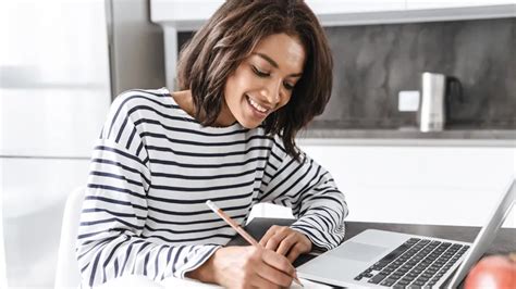 A woman smiling while writing in a notebook and using a laptop at home.