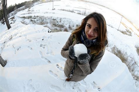 A young and joyful Caucasian girl in a brown coat holds a snowball in ...