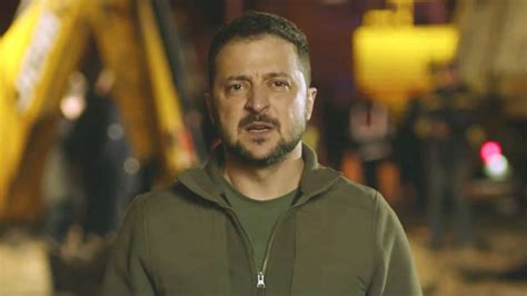 Zelensky wearing a green shirt speaks in a dimly-lit room. An out-of-focus Christmas tree is in the