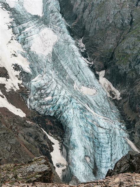 Vertical mountain landscape with blue long vertical glacier tongue with cracks among rocks ...