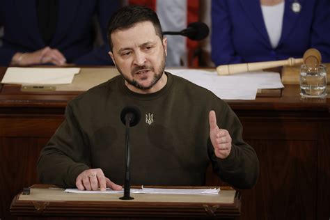 Zelensky wearing a green shirt speaks in a dimly-lit room. An out-of-focus Christmas tree is in the