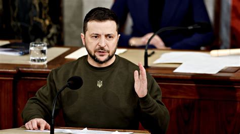 Zelensky wearing a green shirt speaks in a dimly-lit room. An out-of-focus Christmas tree is in the