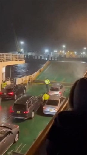 Seattle Ferry has disaster Port of Seattle — nighttime winter storm at a busy ferry terminal. Handheld phone footage from an upper passenger deck shows a powerful wave slam against the side loading bay, forcing seawater across the lower vehicle deck. Multiple cars and SUVs begin sliding more aggressively across the slick surface, bumpers tapping and alarms blaring as crew in reflective rain gear struggle to brace themselves and signal drivers to stay inside. Wind roars, metal creaks, and passeng