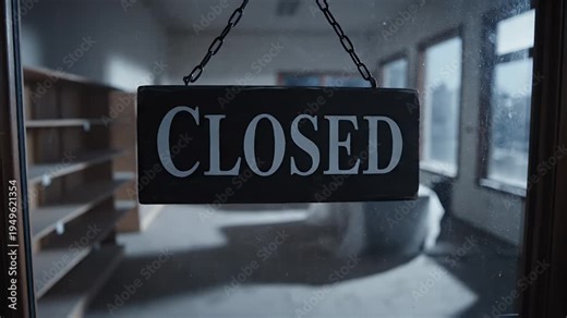 Hand flipping closed sign on a store door in a dimly lit room with bookshelves and windows displaying a winter scene signifying business closure and end of operations