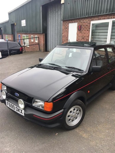 FORD FIESTA XR2 This car is a Ford Fiesta XR2 (second generation, commonly known as Mk2), a classic British hot hatch produced between 1984 and 1989. Exterior Features • Color and Styling: Finished in glossy black paint with distinctive red pinstripes running along the sides and around the wheel arches, a signature look for the XR2 model (though factory colors varied; black with red accents was a popular combination, often seen on well-preserved or restored examples). • Body Kit: It features the