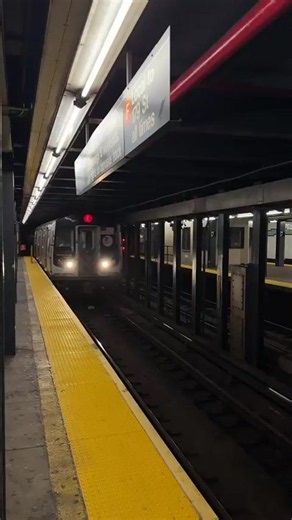 A train approaches a subway station platform. The train is silver with windows that show blurry reflections and silhouettes of passengers. The platform has yellow safety strips along the edge. A sign above the platform indicates the train is the F train, local to 179 St, all times. Fluorescent lights illuminate the station ceiling. The train is moving from right to left in the frame and blurring as it passes. in New York, United States of America | Collective Memory