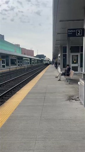A train approaches a station platform. The train is green and white. A person is sitting on a bench on the platform, looking at their phone. The platform has yellow tactile paving along the edge. The sky is overcast. There is a sign that says "Platform 2". in Toronto, Canada | Collective Memory
