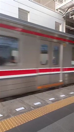 A train moves past a station platform. The train is silver with red stripes. The platform is grey with yellow tactile paving and white markings. Two people are standing on the platform, waiting. One person wears a yellow shirt and a backpack. The other person wears a dark shirt and a hat. in East Jakarta, Indonesia | Collective Memory