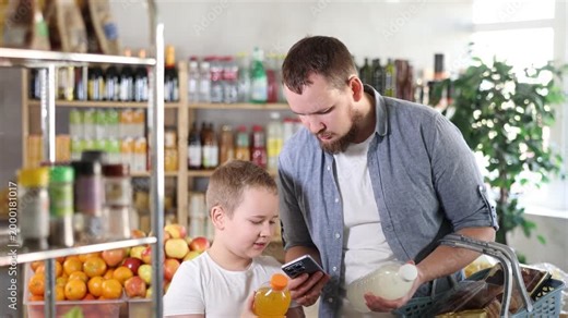 Positive young man with interested preteen son making purchases in local grocery store, using smartphone to scan barcodes on colorful bottles of sweet drinks for online payment