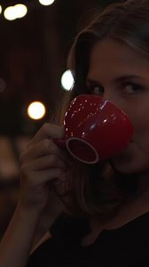 Beautiful young female at cafe drinking coffee or tea, looking into the camera and smiling. Media. Portrait of a flirting woman drinking hot beverage at night summer cafe.