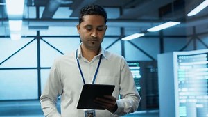 Programmer walking in high tech data center used for managing infrastructure energy consumption, working on tablet. Indian man using device in workspace used for automation in server farm, camera A