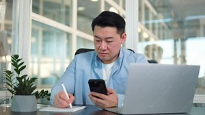 Busy communications department worker using cell phone and copying information into notepad in bright corporate office. Responsible negotiator saving partner's contact data from electronic device.