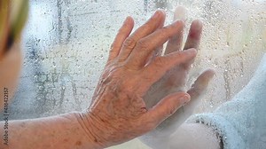 Old hands on the window pane close-up. An old grandmother communicates through a window with loved ones during the coronavirus pandemic. An old and a young hand touching through the glass.