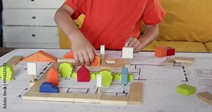 Little child drives wooden car figurine along block track on architectural blueprint closeup. Preschooler boy plays with toy town and auto at table