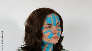 Young woman applied light blue kinesio strips taping face, forehead, neck on white background. Fight against skin aging and wrinkles. Physiotherapy, kinesiology and recovery treatment