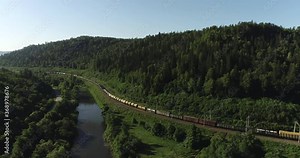 Freight train with cargo carriages an electric locomotive by two-sided Siberian railways near river in the forest mountains / Aerial drone mid view at summer sunset