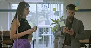 Two colleagues brainstorm and write ideas on a glass board with sticky notes in a modern office, fostering creativity and collaboration.