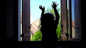 Silhouette of a little girl jumping on the bed on the background of an open window early in the morning