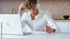 Young adult watching video course and making notes. Student wearing headphones using laptop on his kitchen for learning from online video course and writing down information. Handheld shot