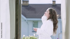 Beautiful woman enjoying sunny morning with cup of coffee on balcony. Video. Attractive woman in white coat is relaxing in expensive hotel and enjoying sunny morning