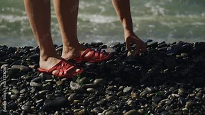 A girl picks up a pebble from the beach while walking along the seashore. On the girl's feet are red flip-flops.